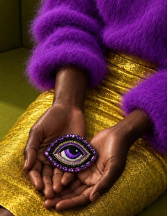 Gros plan sur des mains présentant la broche Œil Céleste violette sur une jupe dorée